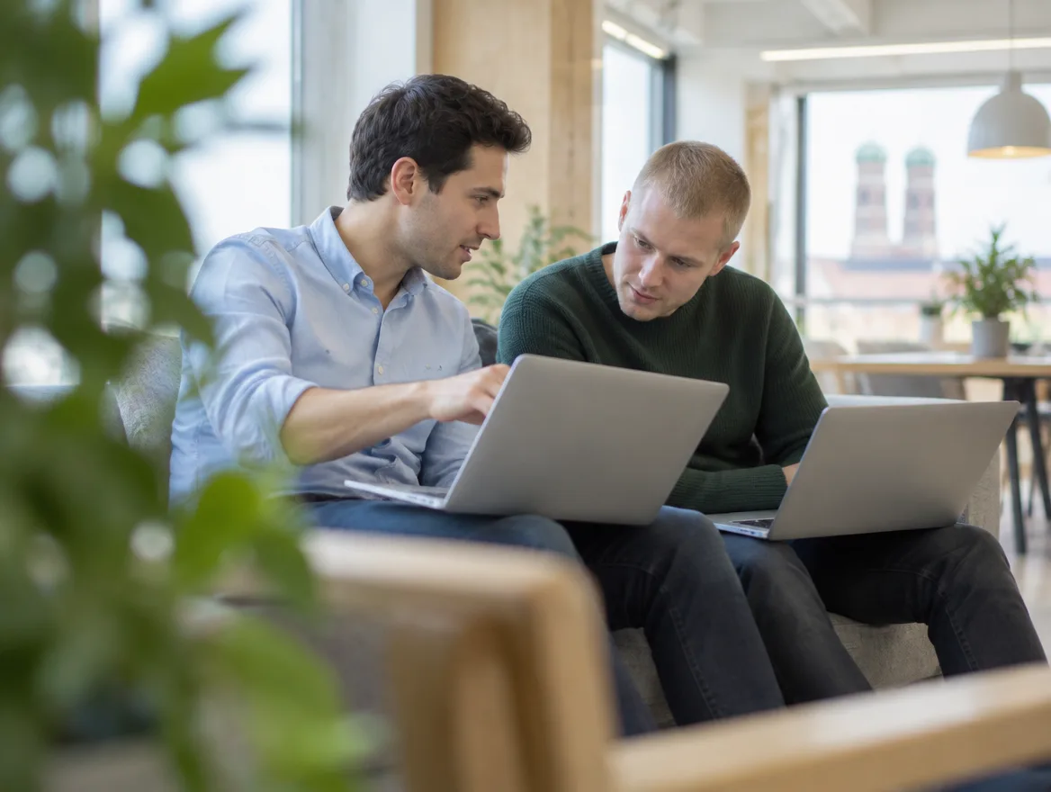 Zwei Engineers sitzen mit Laptops auf einem Sofa, einer dreht seinen Bildschirm zum anderen und erklärt